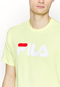 Dzeltena T-krekls ar īsām piedurknēm, kurā ir izcils balts "FILA" logo ar sarkano akcentu, izgatavots no viegla kokvilnas materiāla.