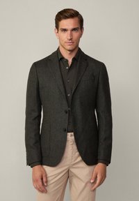 Blazer grigio scuro realizzato in tessuto testurizzato, con una chiusura a due bottoni, rever a scollo e tasche laterali. Indossato sopra una camicia nera.