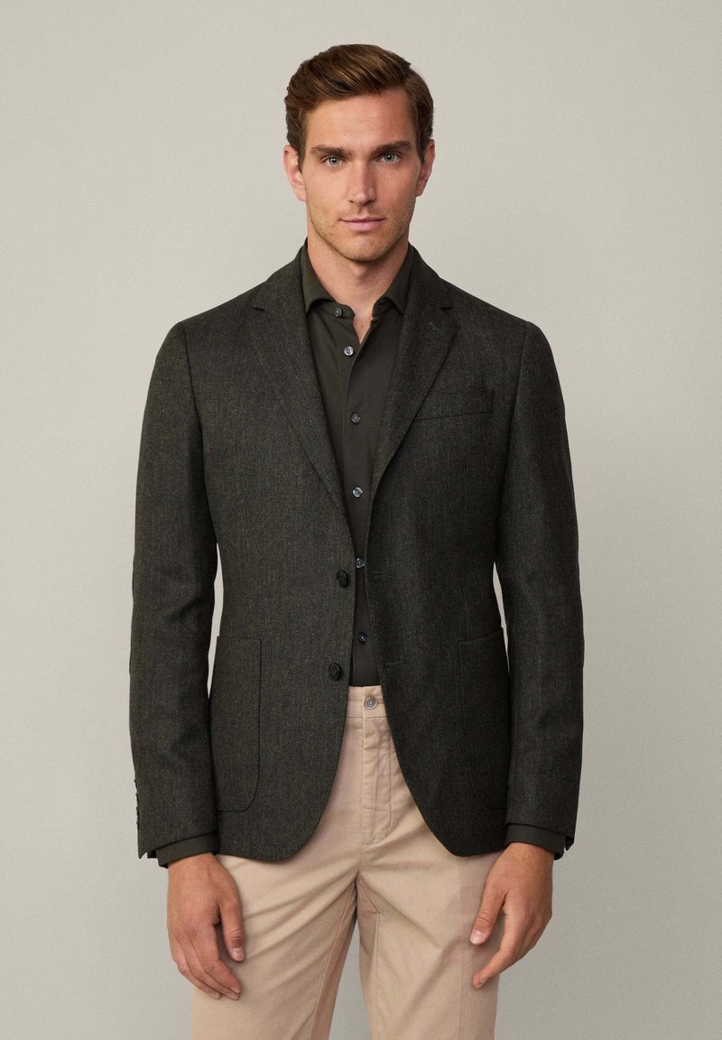 Blazer grigio scuro realizzato in tessuto testurizzato, con una chiusura a due bottoni, rever a scollo e tasche laterali. Indossato sopra una camicia nera.