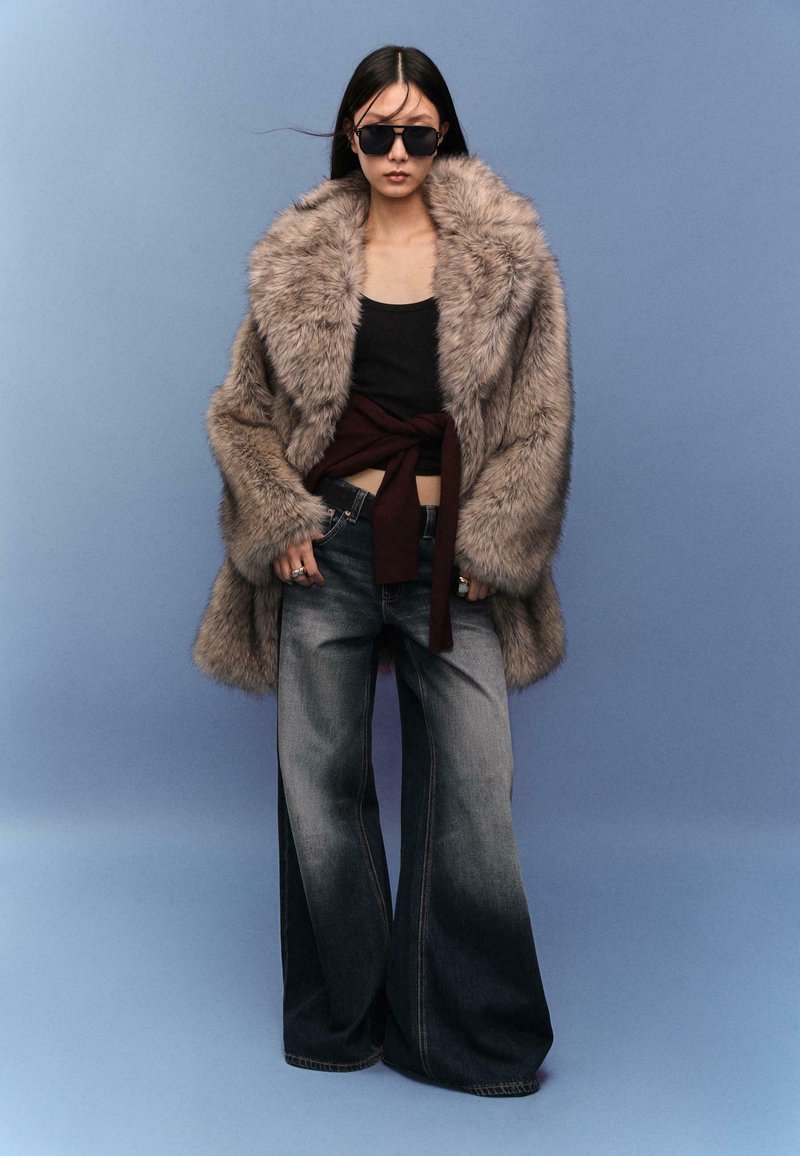 Cappotto in pelliccia sintetica di colore marrone chiaro, top nero, jeans scuri a gamba larga, tessuto borgogna legato in vita, occhiali da sole neri. Sfondo blu.