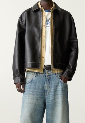 Homme portant des vêtements superposés avec une veste en cuir noire, une chemise à carreaux jaune, un t-shirt blanc graphique et un jean bleu clair avec les mains dans les poches.