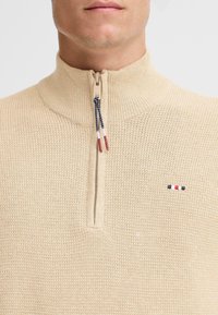 Bela pletena jopica z visokim ovratnikom, polovičnim zipom in teksturirano tkanino. Ima majhen vezen logotip v rdeči, beli in modri barvi.