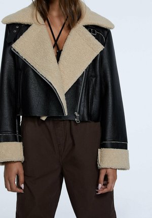 Vrouw draagt een zwarte leren jas met beige shearlingkraag en manchetten, gecombineerd met een bruine broek, zichtbaar van schouders tot heupen.