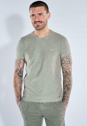 Mann mit kurzen Haaren und Tätowierungen auf beiden Armen, trägt ein hellgrünes T-Shirt und passende Hosen, steht mit den Händen in den Taschen.