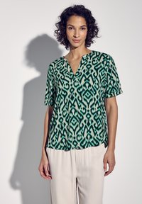 Blusa a maniche corte verde e bianca con scollatura a V, caratterizzata da un design astratto e realizzata in un tessuto leggero e liscio.