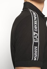 Svart polo-skjorta i bomull med krage och korta ärmar. Märkbart vit logoband längs axlarna och ärmarna.