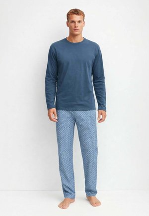 Uomo in piedi a piedi nudi che indossa una camicia blu scuro a maniche lunghe e pantaloni del pigiama azzurri con fantasia, su sfondo bianco semplice.