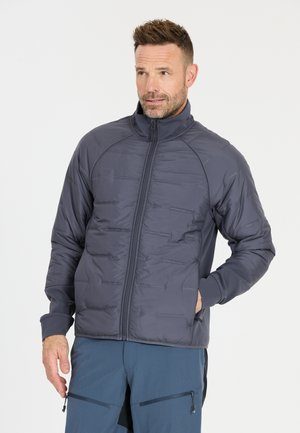 Mann trägt eine marineblaue gesteppte Jacke mit Reißverschluss und hohem Kragen sowie blaue Reißverschluss-Hose, hält eine Hand in der Jackentasche und posiert.