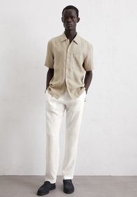 Camicia a maniche corte a righe marrone chiaro con colletto, abbinata a pantaloni larghi in lino bianchi e scarpe nere senza lacci.