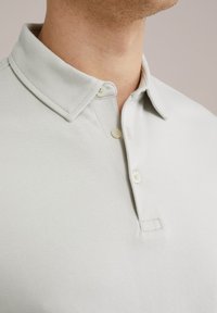 Svetlo siva polo majica s preklopnim ovratnikom, dvema gumboma, mehko teksturo in šivanim detajlom na ovratniku in gumbni ploskvi.