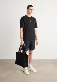 T-shirt nera a maniche corte con logo, pantaloni corti neri con dettagli blu e borsa tote nera. Modello che indossa scarpe da ginnastica bianche, in piedi su un pavimento neutro.