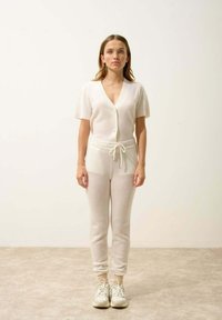 Ljus beige stickad tvådelad outfit med kortärmad cardigan och byxor med dragsko. Mjuk textur, figurnära form och elastiska manschetter vid anklarna.