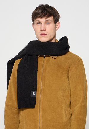 Uomo che indossa una giacca in camoscio color cuoio e una sciarpa nera lavorata a maglia avvolta intorno al collo, su uno sfondo chiaro e uniforme.