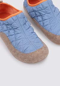 Zapatillas interiores acolchadas de color azul con una suave suela de lana en tono marrón claro y un forro interior naranja. Presentan una parte superior fruncida y una puntera redondeada.