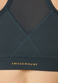 Tempererat svart sportbehå med en texturerad meshrygg. Har en korsad design och "SWEDMOUNT"-logotyp i guld vid midjan.