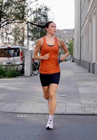 Løbetræningsudstyr: orange ærmeløs top, sorte shorts, mønstrede hvide sokker. Urbant miljø med bygninger, træer og parkerede cykler.
