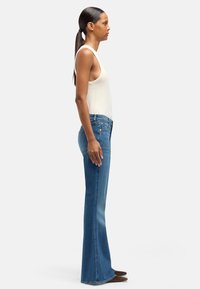 Jasnoniebieskie dzwony jeansowe z wyblakłym odcieniem, pięcioma kieszeniami i małymi metalowymi dodatkami. W połączeniu z dopasowanym białym topem na ramiączkach. Widok z boku.