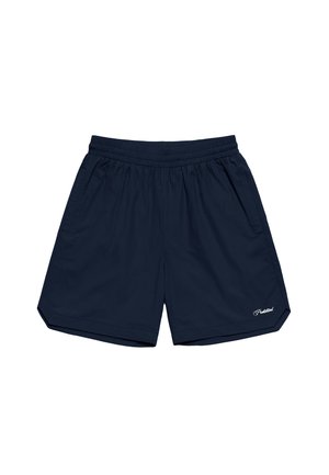 Mand iført navyblå shorts, hvide sokker med sort tekst og sorte og hvide loafers, stående foran en ensfarvet baggrund.