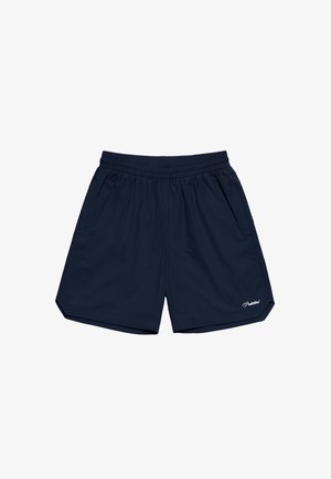 Mann trägt marineblaue Shorts, weiße Socken mit schwarzem Text und schwarz-weiße Loafers, steht vor einem einfachen Hintergrund.