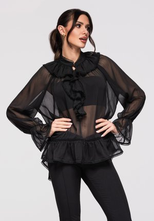 Femme portant un chemisier noir transparent avec col à volants, manches longues bouffantes et bas à basque, associé à un pantalon noir ajusté.