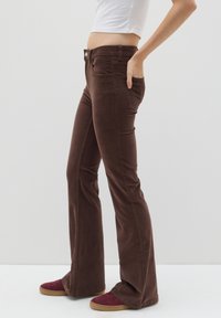 Bruine corduroy flared broek met losse zakken aan de voorkant, een getailleerde taille en een lichte glans. Gecombineerd met maroon sneakers op een witte achtergrond.