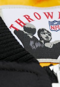 Schwarzer Stoff mit strukturiertem Muster und gelben Nähten; verfügt über ein grafisches Etikett mit dem Text "THROW" und dem NFL-Logo, das stilisierte Figuren zeigt.