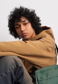 Brun hættetrøje, grøn taske med lynlås, denim jeans. Personen har krøllet hår, septum piercing og sidder op ad en neutral baggrund.