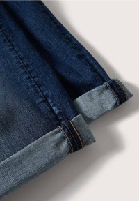 Jean en denim bleu foncé avec les revers retroussés, dévoilant le tissu intérieur plus clair et les coutures oranges le long des coutures.