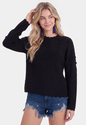 Lyshåret kvinde iført en sort tekstureret sweater med pomponer og blå kvastøreringe, kombineret med slidte denimshorts.