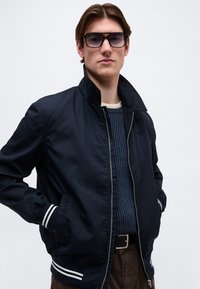 Jeune homme portant une veste noire zippée avec des rayures blanches aux poignets et à l'ourlet, un pull côtelé bleu, un pantalon marron et des lunettes de soleil foncées.