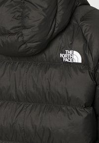 Doudoune noire matelassée horizontalement avec une capuche, arborant le logo blanc "The North Face" sur le haut du dos côté droit.