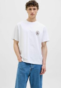 Hvid bomulds t-shirt med korte ærmer og rund halsudskæring, prydet med et cirkulært grafisk design på venstre bryst, stylet med blå denim jeans.