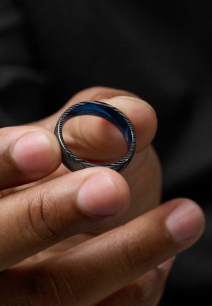 Black metal ring with a matte finish and a textured edge, featuring a blue interior and a smooth surface. Held between fingers.