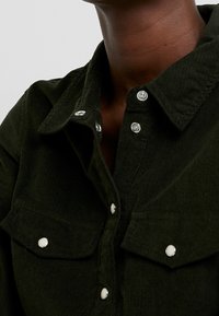 Mörkgrön kordflorse skjorta med krage, silverfärgade tryckknappar och framfickor på bröstet. Texturen är ribbad och mjuk.