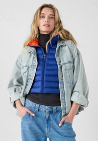 Chaqueta vaquera de lavado claro, superpuesta sobre un chaleco acolchado azul con ribete de cuello naranja, complementada con un cuello alto gris y vaqueros azules.