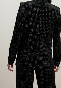 Blazer en velours noir à pois blancs. Présente une coupe ajustée, des manches longues et une texture lisse. La vue de dos met en évidence les détails de couture.
