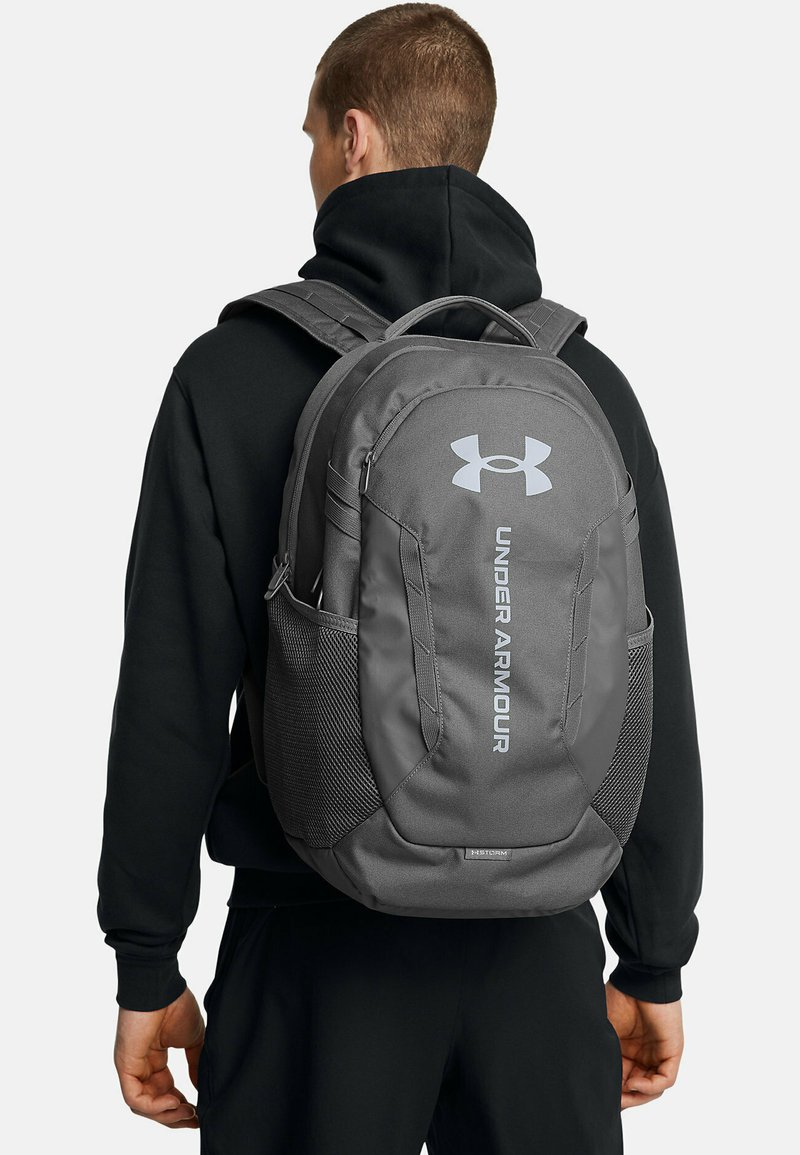 Zaino grigio con logo Under Armour, spallacci imbottiti, tasche laterali in rete e un esterno resistente e testurizzato. Adatto per lo sport o l'uso quotidiano.