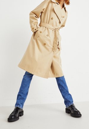 Vrouw met een beige trenchcoat, blauwe spijkerbroek en zwarte loafers, staand met wijd gespreide benen tegen een effen witte achtergrond.