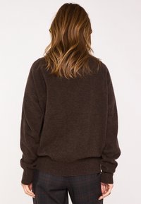 Pull en maille marron avec une coupe ample, des manches surdimensionnées et un ourlet en côtes. Porté sur un pantalon à carreaux, mettant en avant une texture douce.