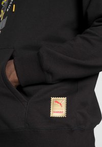 Sort sweatshirt med kengurulomme, ribbede manchetter og et guldmærke med rødt Puma-logo og "HARIBO" tekst nederst til venstre.