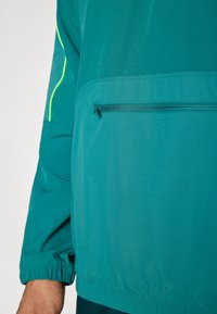 Τζελ ανθεκτικό μπουφάν με υφή, που διαθέτει τσέπη με φερμουάρ και ελαστικές μανσέτες για σταθερή εφαρμογή. Ξεχωρίζουν λεπτομέρειες σε νέον κίτρινο χρώμα.