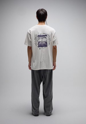 Persoon staat met de rug naar de kijker, draagt een lichtgrijs T-shirt met een abstract paars ontwerp en donker, loszittend broek tegen een effen achtergrond.