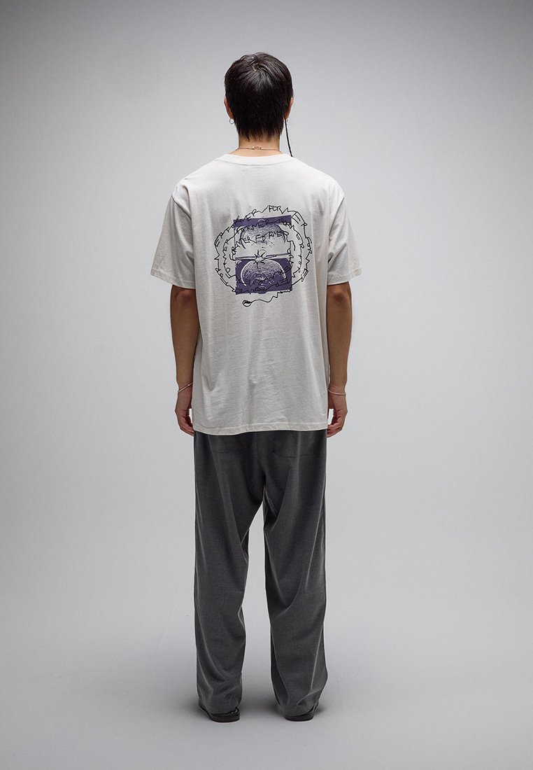 Άτομο στέκεται πλάτη, φοράει ανοιχτό γκρι μπλουζάκι με αφηρημένο μωβ σχέδιο και σκούρο φαρδύ παντελόνι, μπροστά σε απλό φόντο.