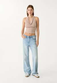Beige halterneck crop top med draperad halsringning, parat med ljusblå vida jeans. Modellen har på sig silverfärgade sneakers och står med händerna i fickorna.