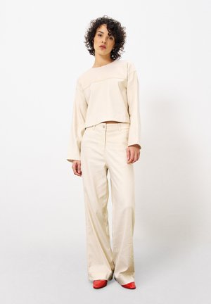 Beige verkürzter Pullover mit einer horizontalen Naht; kombiniert mit weit geschnittenen beigen Hosen. Das Model trägt rote spitze Schuhe und steht vor einem hellen Hintergrund.