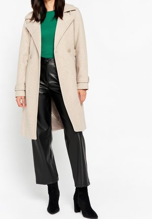 Kobieta ubrana w beżowy długi płaszcz, zielony prążkowany sweter, czarne spodnie z imitacji skóry oraz czarne botki na wysokim obcasie stoi na białym tle.