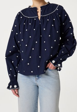 Marineblauwe blouse met lange mouwen, witte bloemen- en hartborduursels, gerimpelde manchetten en een knoopsluiting bij de hals, gedragen met lichtblauwe jeans.