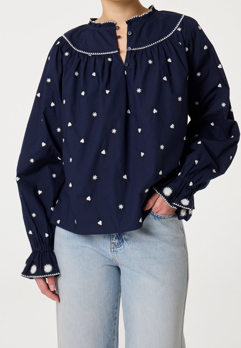 Blouse bleu marine à manches longues avec broderies blanches florales et en forme de cœur, poignets volantés et encolure boutonnée, portée avec un jean bleu clair.
