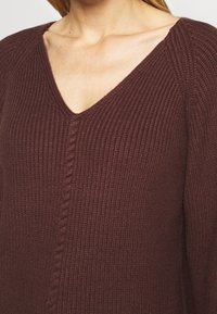 Gros plan sur une femme portant un pull en maille côtelée marron avec un col en V et un détail torsadé central en forme de câble.