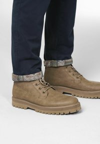 Bruine leren enkellaarzen met een gestructureerd oppervlak, ronde neus en contrasterende rubberen zool. Opgestropen marineblauwe broek met een patroon aan de zoom.